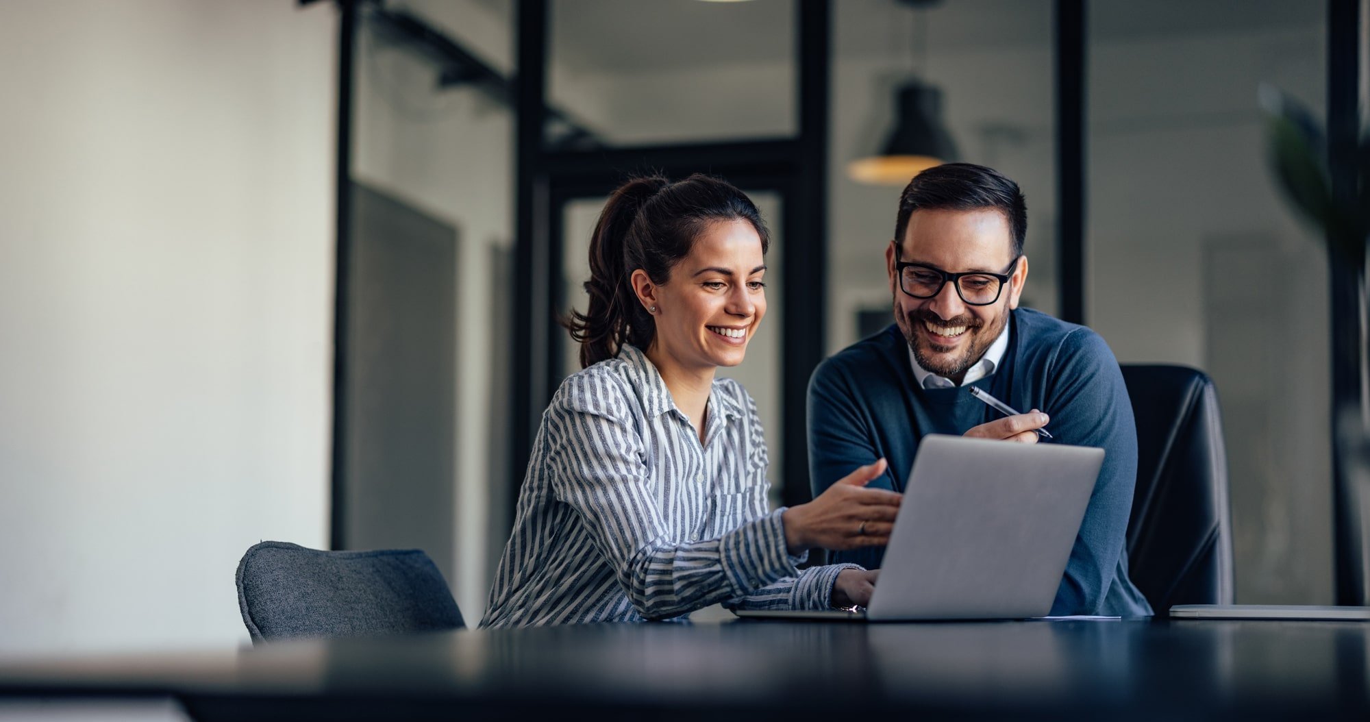Zwei Geschäftspersonen sitzen lächelnd an einem Schreibtisch und schauen gemeinsam auf einen Laptop, während sie über digitale Rechnungsprozesse sprechen.