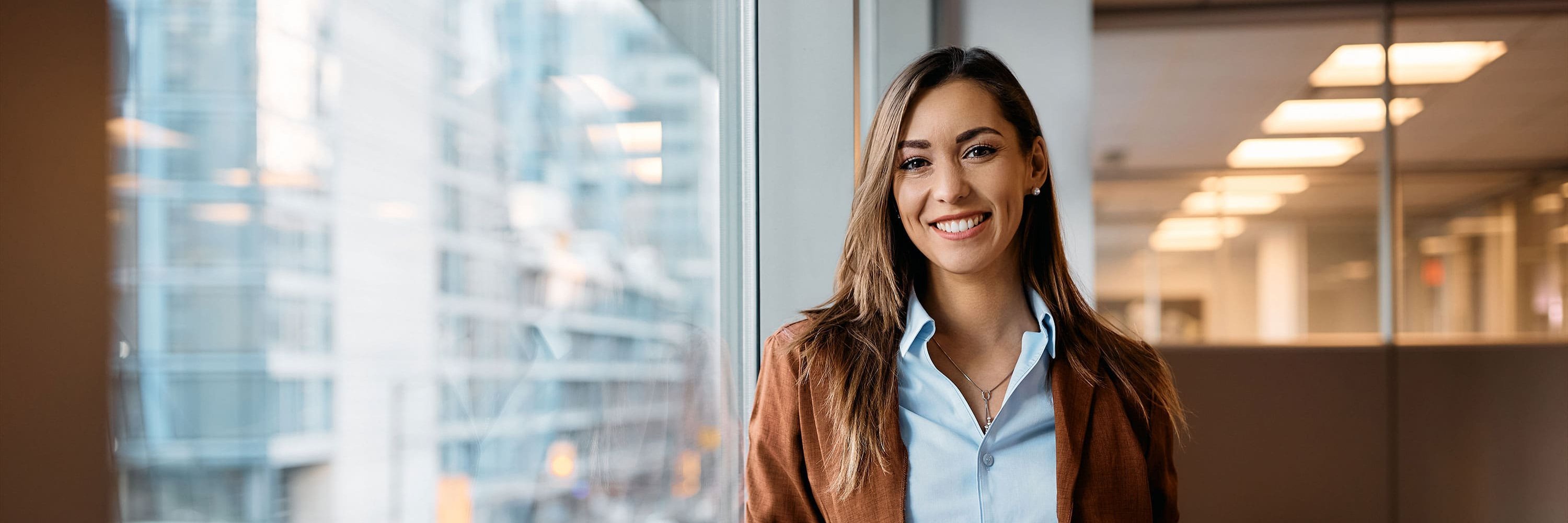 Mitarbeiterin in modernem Büro steht vor großer Fensterscheibe mit Tablet in der Hand und lächelt.
