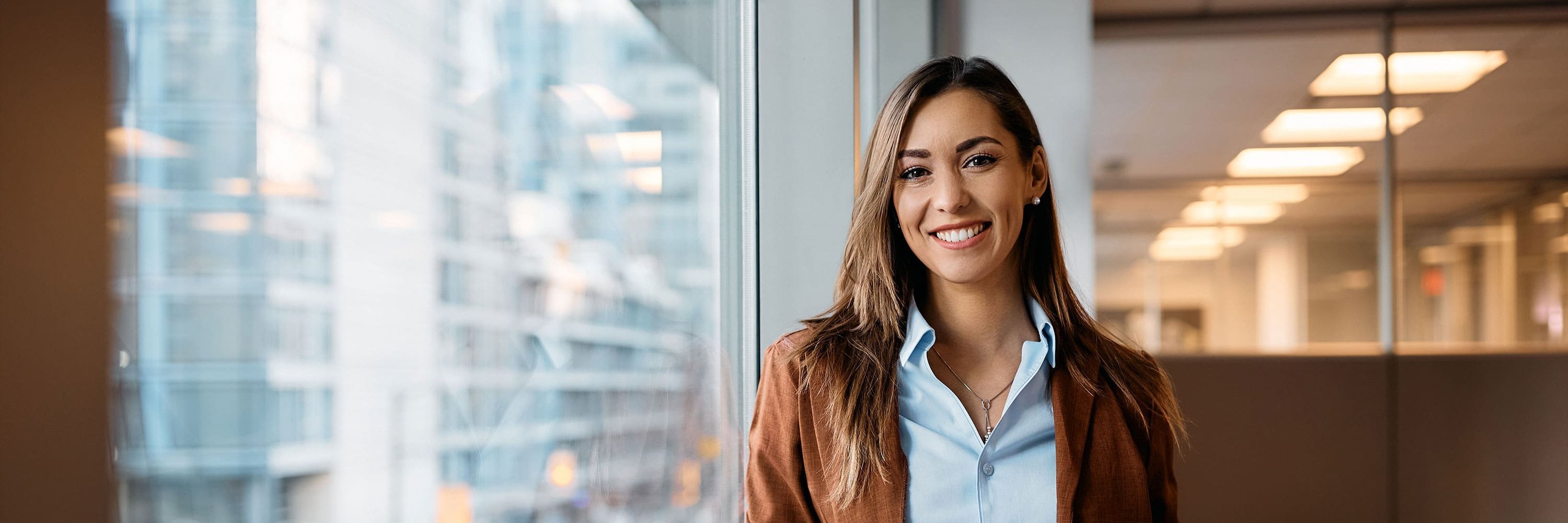 Mitarbeiterin in modernem Büro steht vor großer Fensterscheibe mit Tablet in der Hand und lächelt.