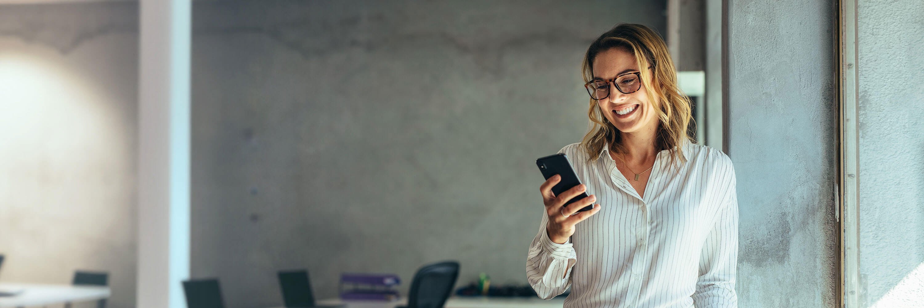 Frau mit Brille und weißer Bluse hält Smartphone in der Hand und lächelt. 