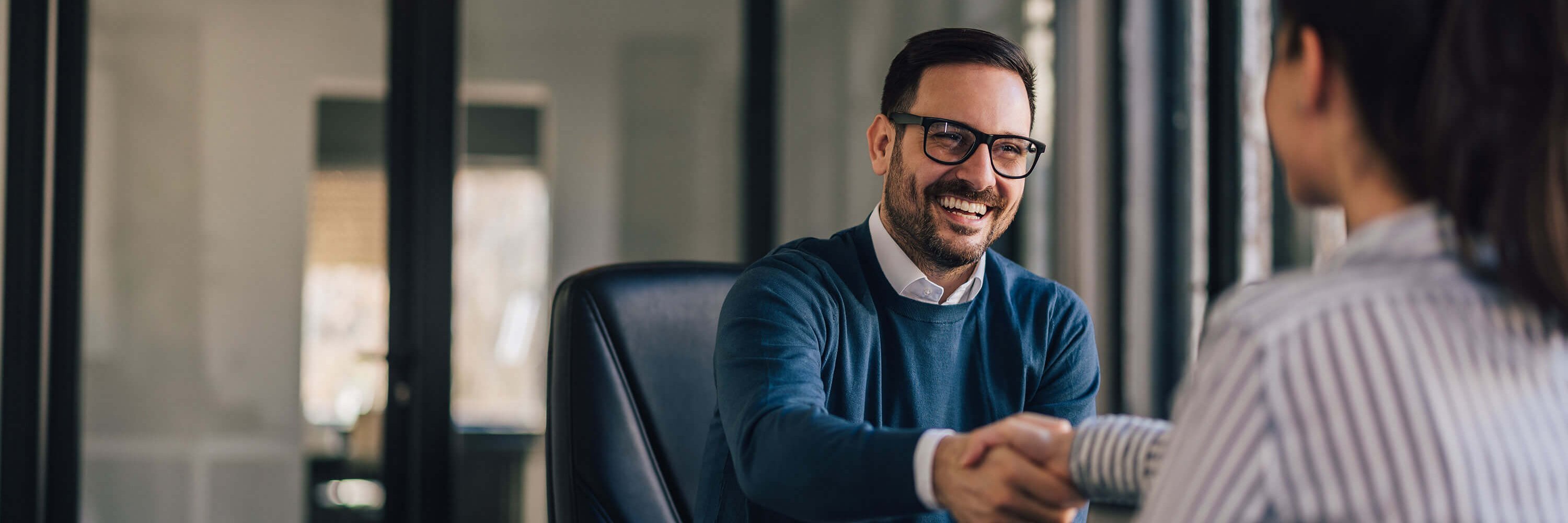 Ein lächelnder Mann mit Brille und eine Frau schütteln sich im Büro die Hände. 