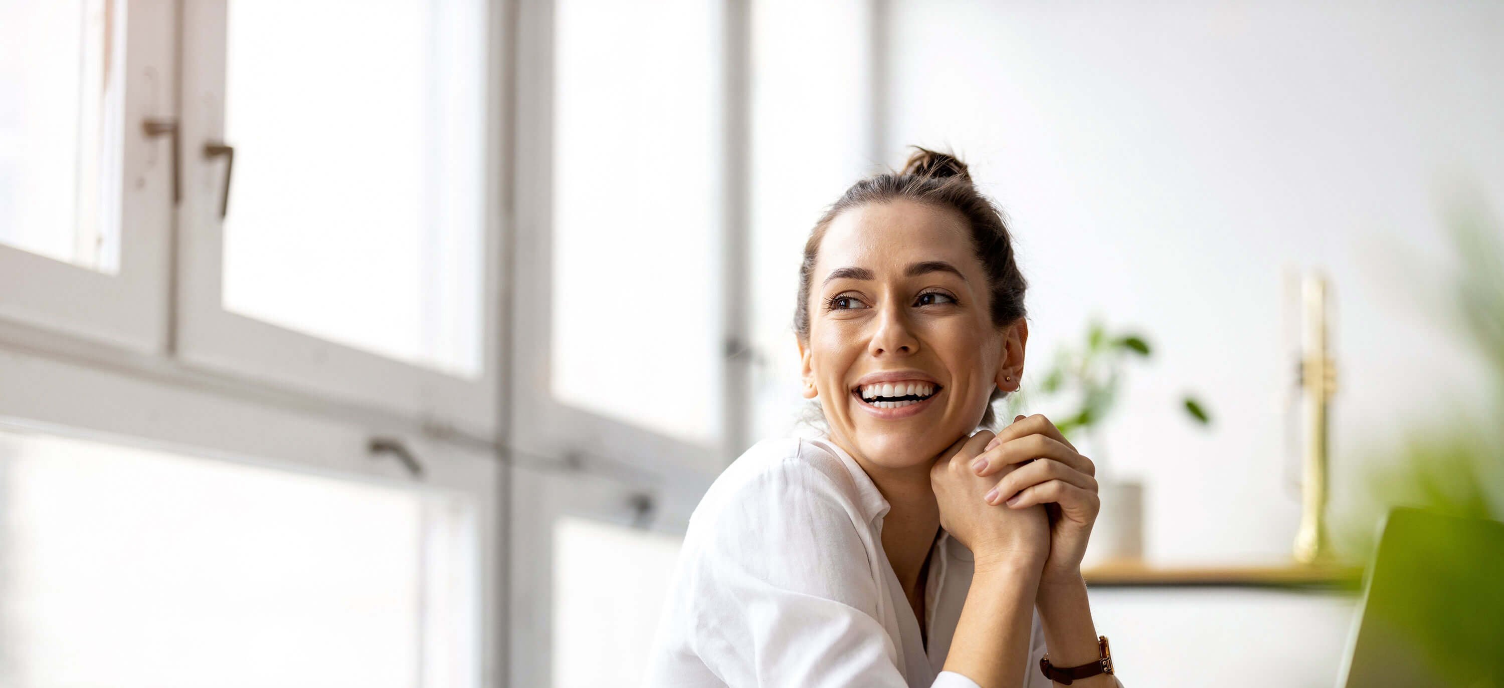 Fröhliche Mitarbeiterin in hellem Büro sitzt von Notebook und schaut lächelnd zur Seite.