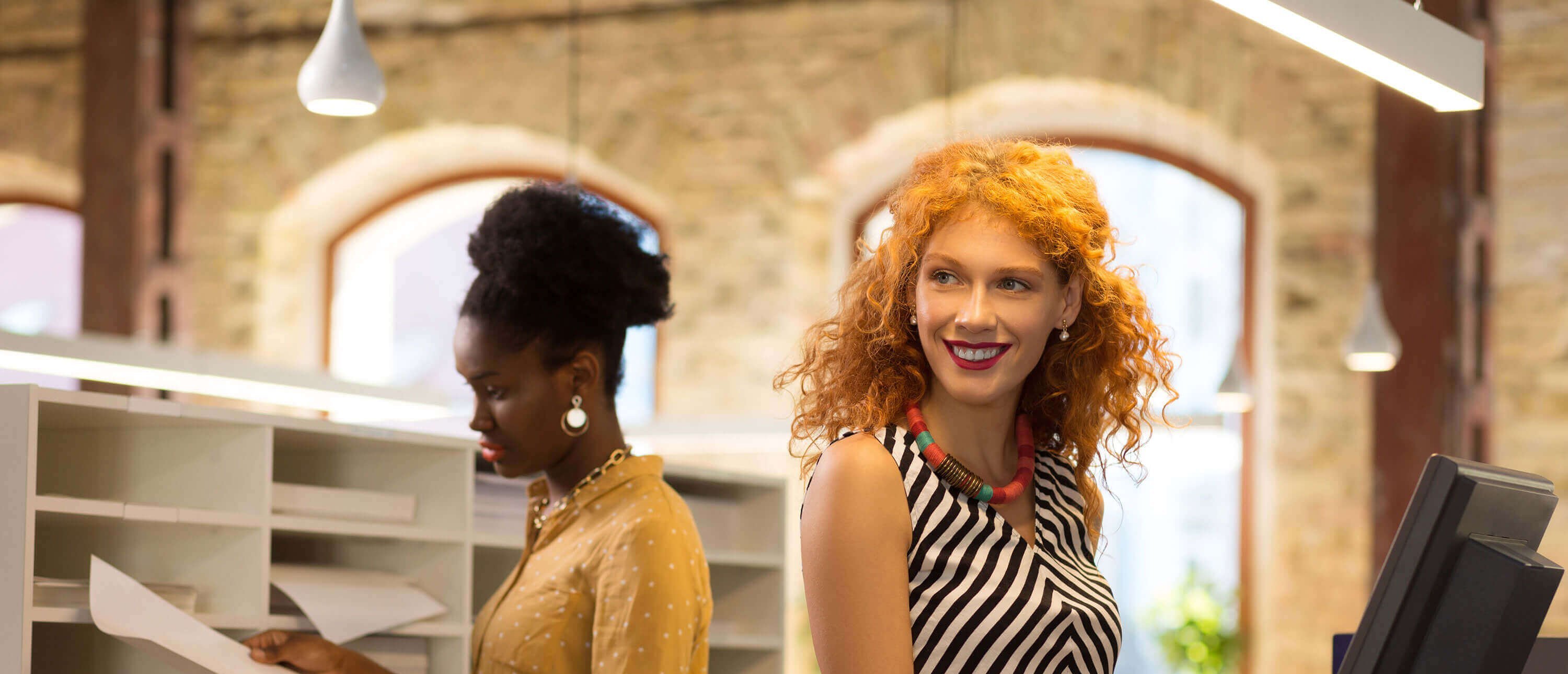 Zwei Frauen im Büro. Eine Frau mit schwarz-weißem Oberteil steht an einem Kassenterminal und lächelt.