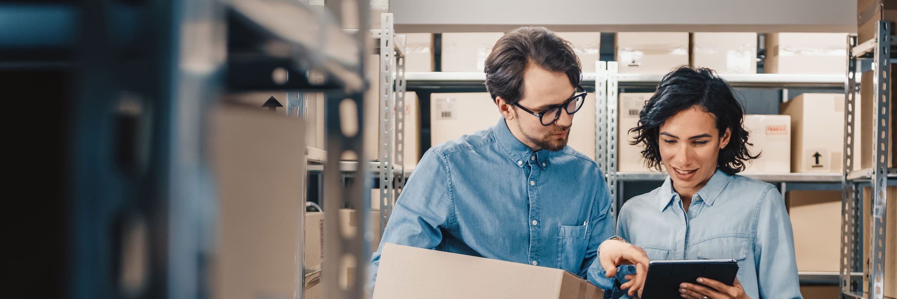 Ein Mann und eine Frau stehen in einem Lager und blicken auf ein Tablet. 