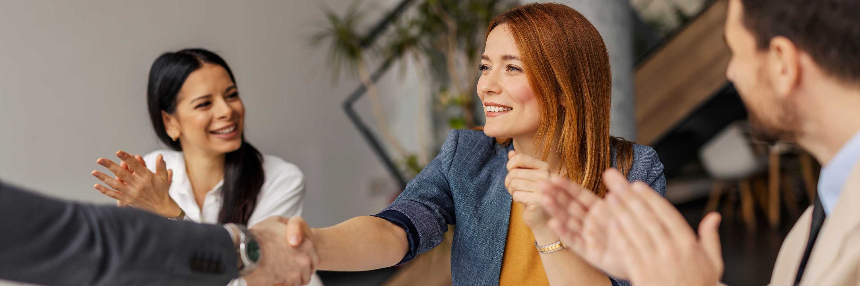 Žena s oranžovými vlasmi s úsmevom podáva ruku mužovi. Iný muž tlieska rukami.