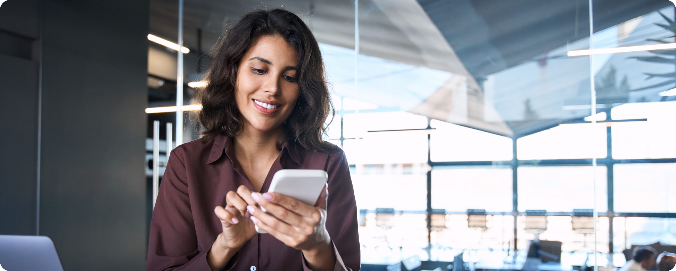 Frau mit braunen Haaren und weinroter Bluse sitzt an Bürotisch und tippt auf ihrem Smartphone. 