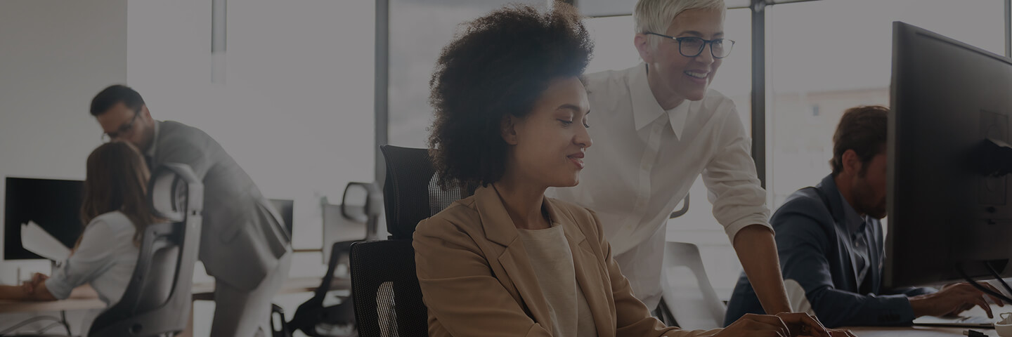 Zwei Frauen im Büro, die auf einen Desktop blicken und lächeln.