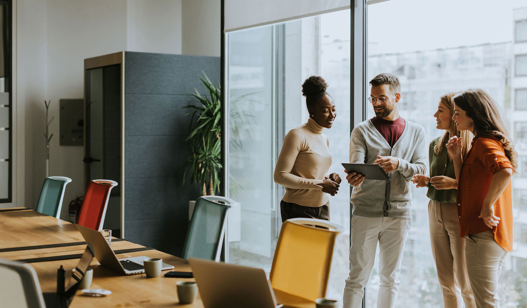 Ein Team von vier Personen steht in einem modernen Büro und bespricht gemeinsam Inhalte auf einem Tablet, umgeben von hellen Farben und natürlichem Licht.