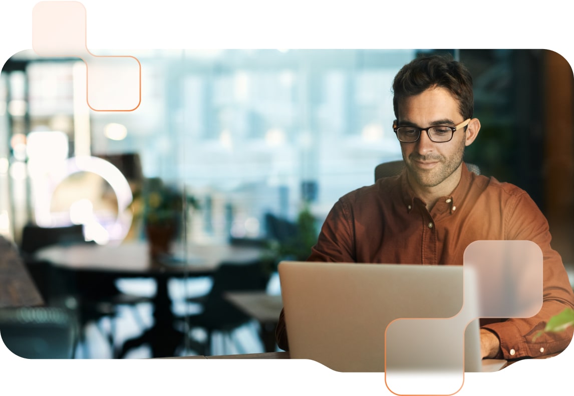 Mitarbeiter sitzt vor dem Notebook-Bildschirm, im Hintergrund ist ein modernes Büro zu sehen, der Mitarbeiter lächelt.