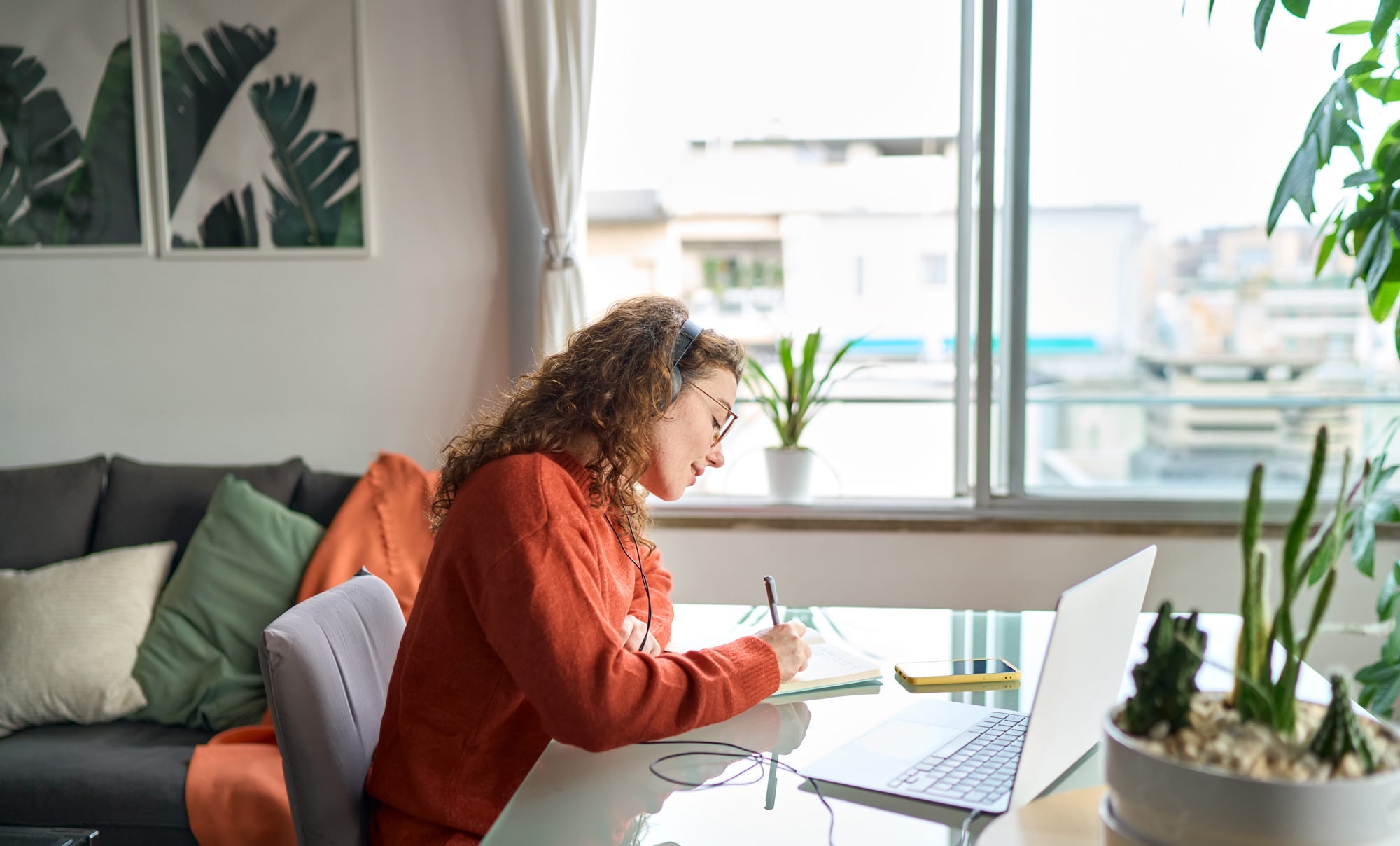   Schülerin lernend im Homeoffice am Schreibtisch mit Notebook, Notizblock, Kopfhörern und Sofa im Hintergrund.