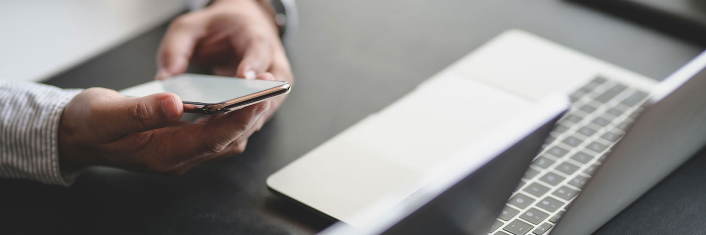 Person, die ein Smartphone in der Hand hält. Vor der Person steht ein Laptop.