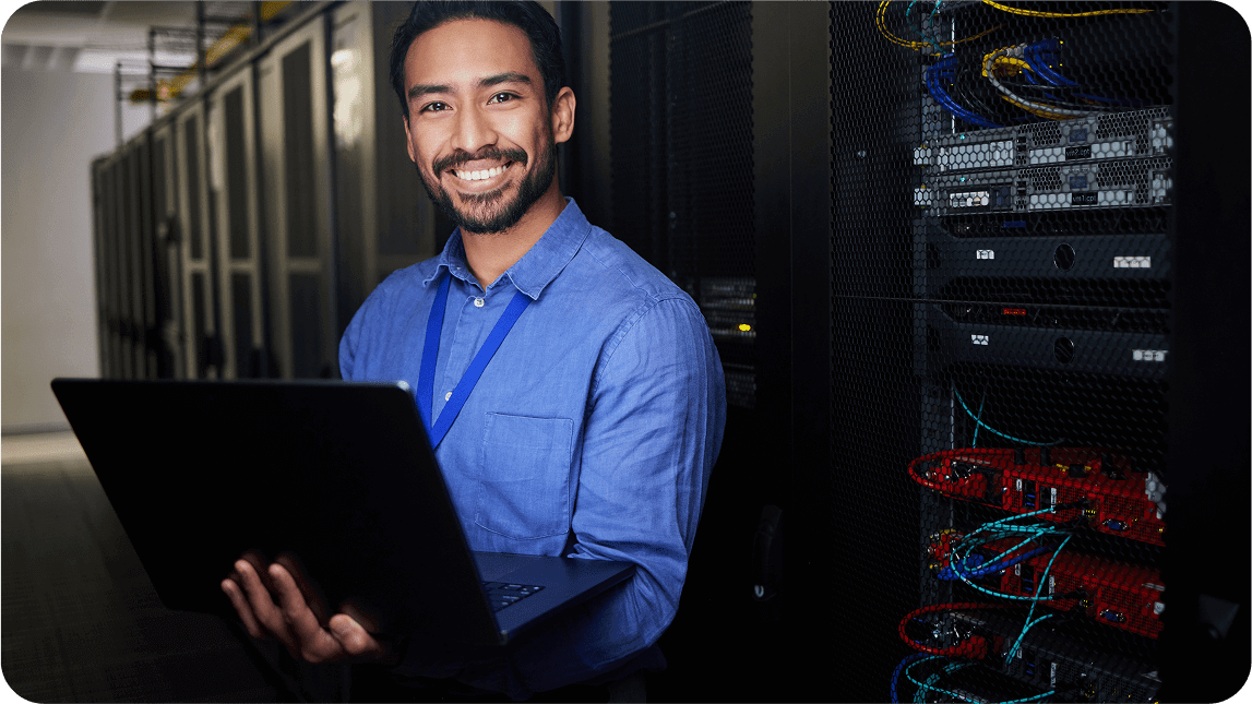 IT Techniker in blauem Hemd mit Notebook im Serverraum.