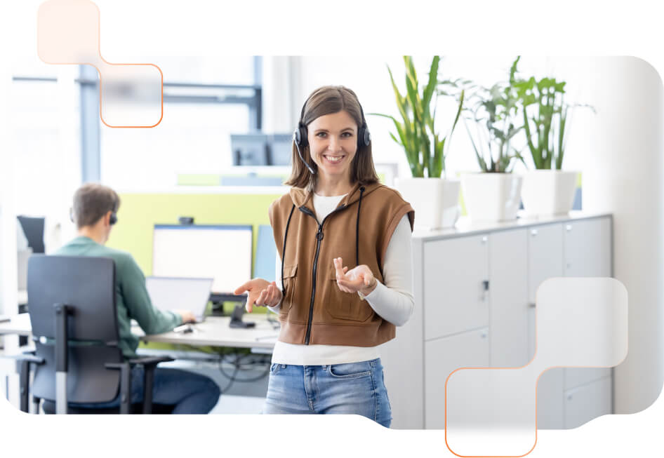 BMD Support Mitarbeiterin BMD Support Mitarbeiterin mit Headset lächelnd und stehend im Büro - im Hintergrund sitzt ein weiterer BMD Mitarbeiter beim PC.