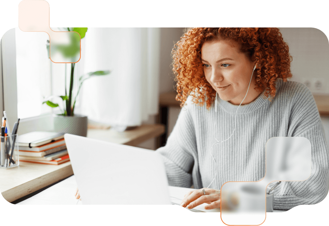Frau mit orangen locken sitzt mit Kopfhörern im Ohr vor einem Laptop und lächelt. 