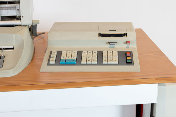 Beige-graue Rechenmaschine mit zwei blauen Tasten und bunten Knöpfen auf Holztisch