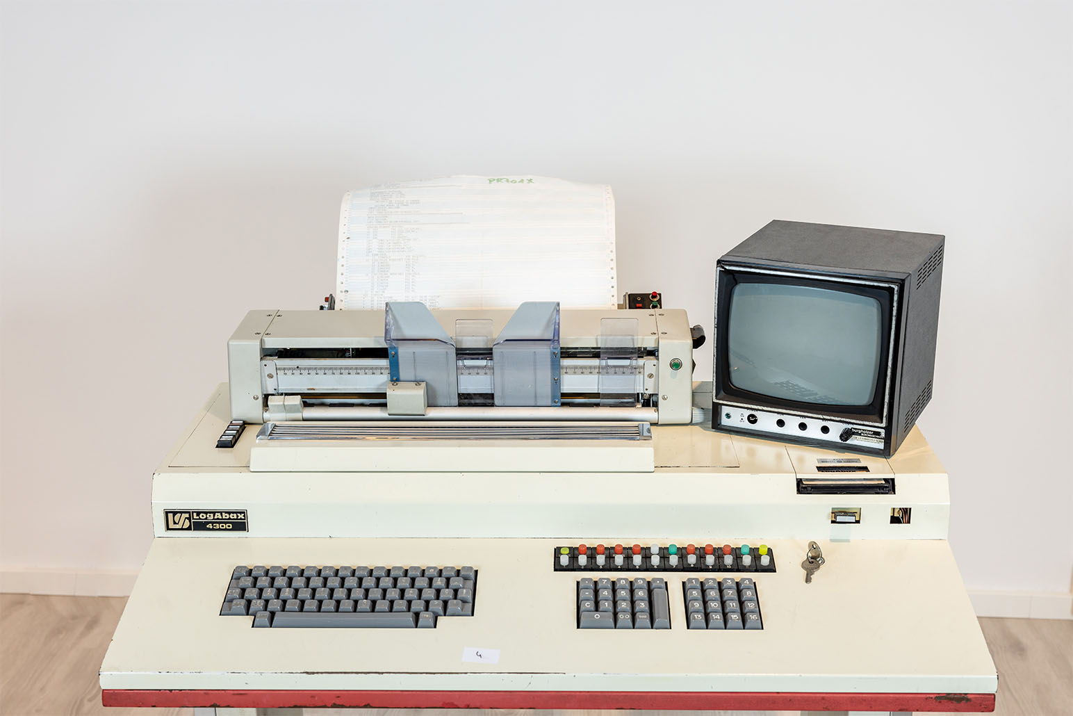 Weißer Computer mit sehr kleinem schwarzen Monitor, grauen Tasten und rotem Streifen vor weißer Wand