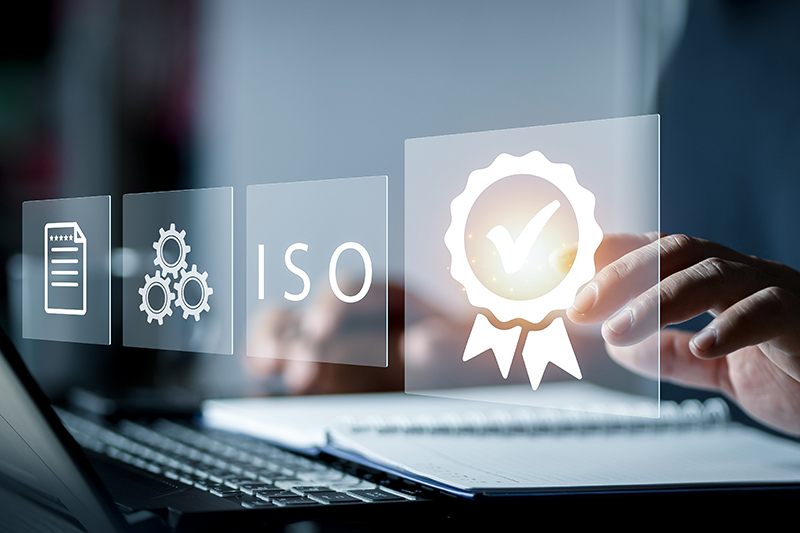 Hände einer Person, die am Notebook sitzt und tippt. Im Vordergrund sind verschiedene Icons zum Thema ISO Zertifizierung abgebildet. [© Montri - stock.adobe.com]