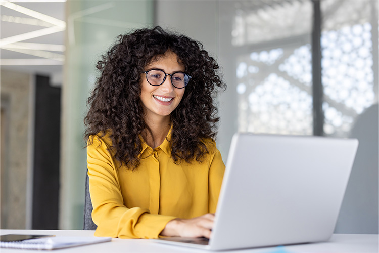 Frau mit schwarzen Locken und Brille tippt lächelnd auf einem Laptop.