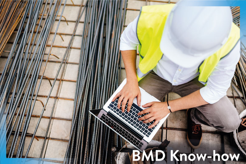 Person mit Leuchtweste, Helm und Laptop sitzt auf einer Baustelle