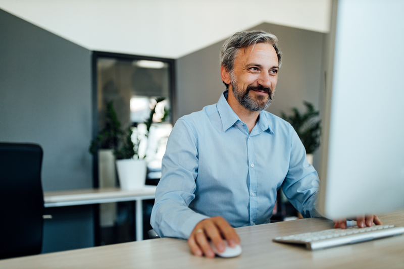 Ein lächelnder Mann mittleren Alters mit Bart und hellblauem Hemd arbeitet an einem Computer in einem modernen Büro.