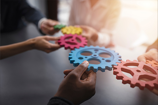 5 színes fogaskerék látható, amelyek egymáshoz kapcsolódnak, és amelyeket 5 kéz tart.