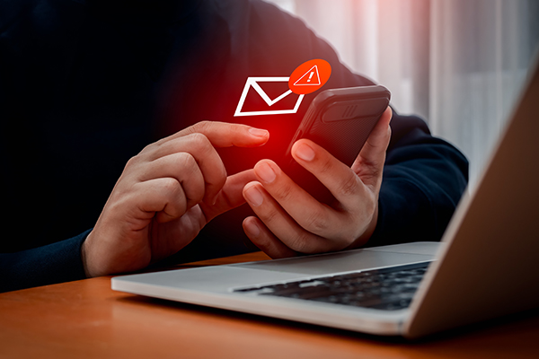 Egy férfi az okostelefonján gépel, mögötte egy laptop áll. Előtte egy hívójellel ellátott betűjel jelenik meg.