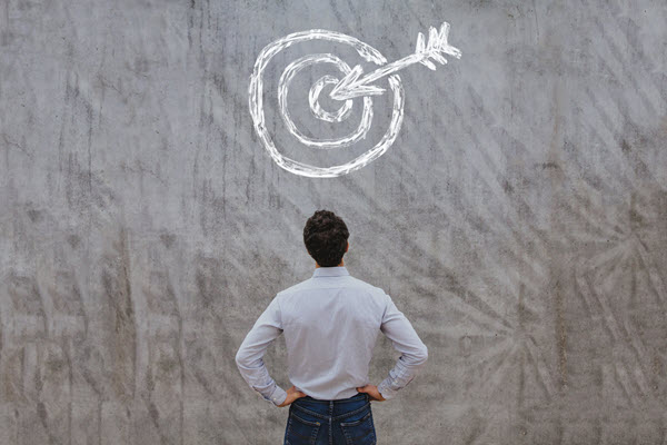 A képen egy nagy fehér céltábla és egy nyílvessző látható, amely eltalálja azt. Egy férfi áll előtte és nézi.