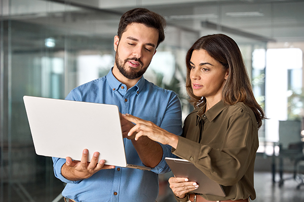 Lát egy nőt egy táblagéppel a kezében és egy férfit egy laptoppal az oldalán, mindketten a képernyőre mutatnak az ujjukkal.