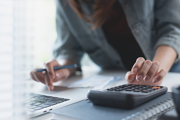 Egy nő egy számológépbe gépel, másik keze egy laptop billentyűzetén van.