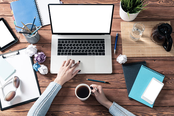 A képen egy nő látható, aki egyik kezével a laptopján dolgozik, a másikkal pedig egy csésze kávét tart.